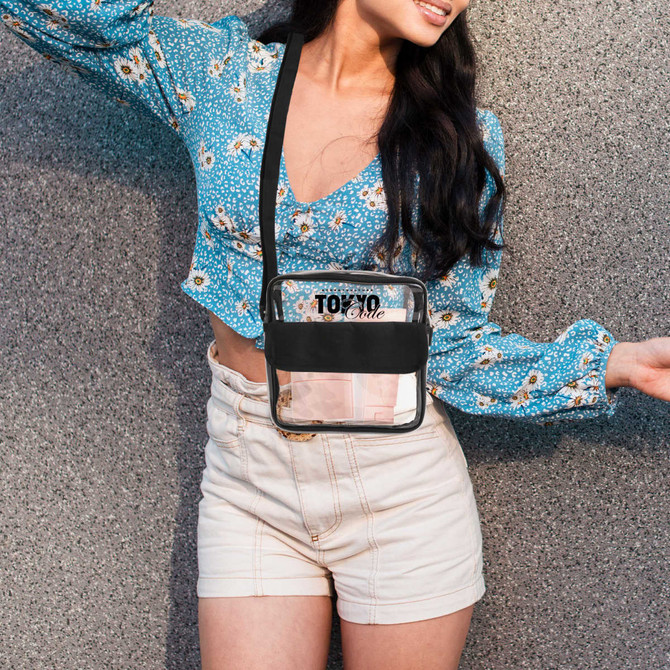A clear crossbody bag with a black trim, worn over a floral blue top and white shorts.