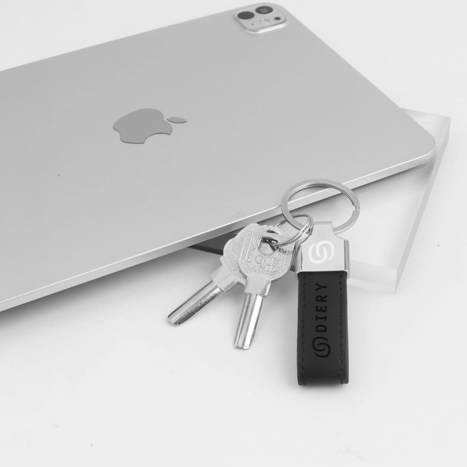 A metal keyring in black PU leather with two silver keys, placed beside a silver tablet. It features a logo.