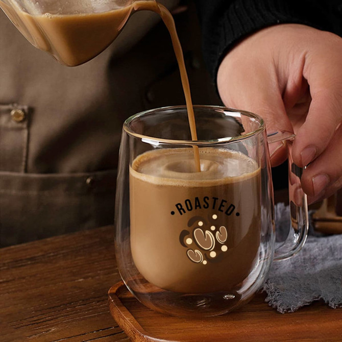 A barrel-shaped double wall mug in clear glass is being filled with a creamy coffee beverage.
