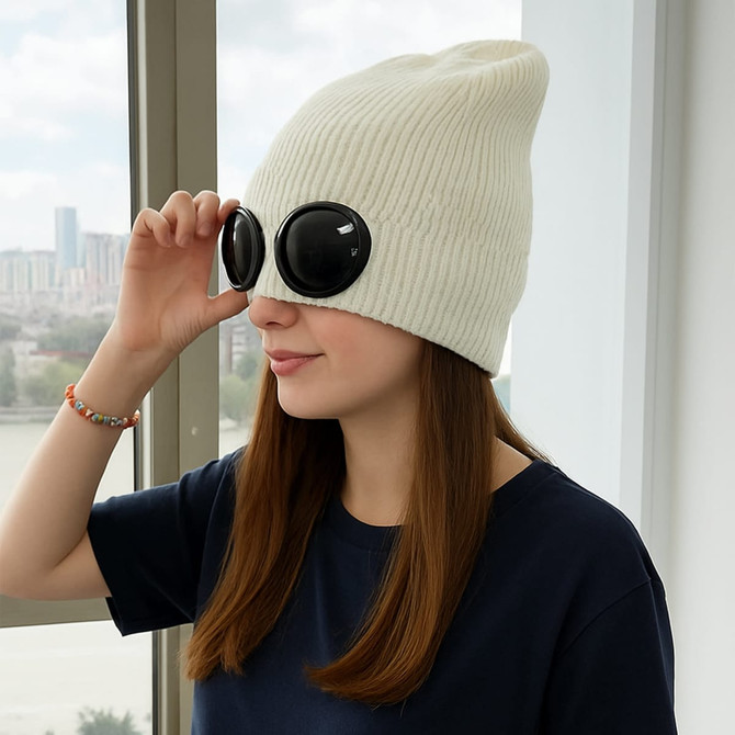 A cream beanie paired with large black goggles, worn by a woman indoors near a window.