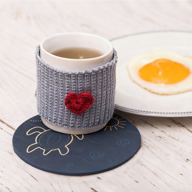 A round soft diatomaceous coaster in dark blue, supporting a cup with a knitted grey sleeve and a heart embellishment.