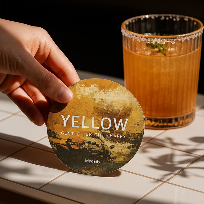A round, frosted metal coaster in yellow with a textured design, held near a glass of cocktail.
