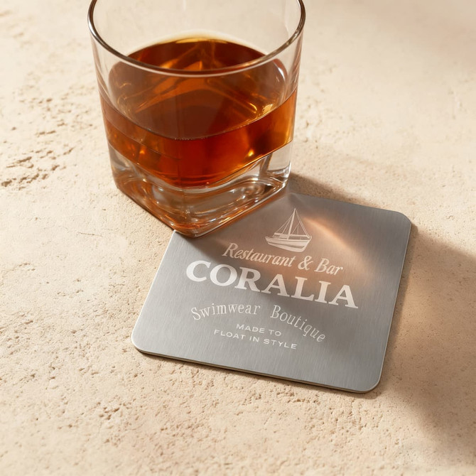 A square metal coaster with a silver finish, placed under a glass of dark beverage. It features a logo.