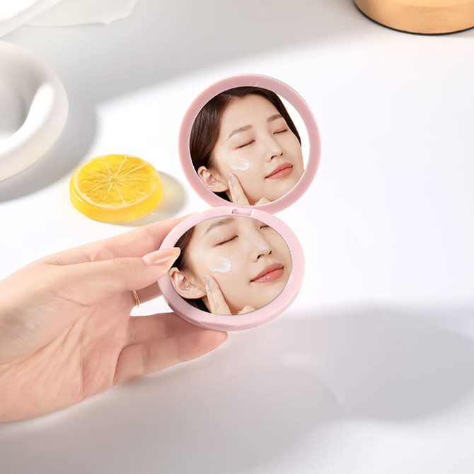 A double-sided round mirror in a pink casing, being held, with a reflection of a person applying skincare.