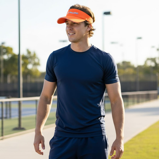 A man wearing a navy athletic shirt and an orange sun visor stands on a tennis court. The scene is sunny and vibrant.