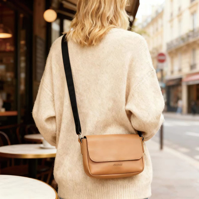 A tan polyurethane shoulder bag with a black strap is worn by a person in a cream sweater.