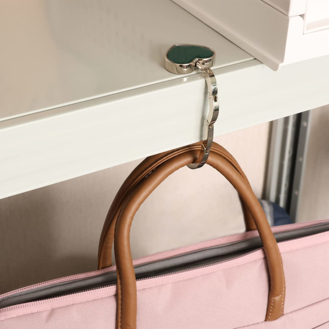 A heart-shaped, silver foldable desk hook is attached to a shelf, holding a pink handbag with brown handles.