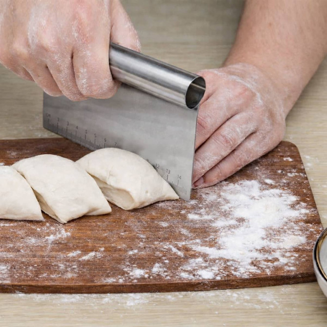 A stainless steel flour cutter with measurements, cutting dough on a wooden board, with flour scattered on the surface.