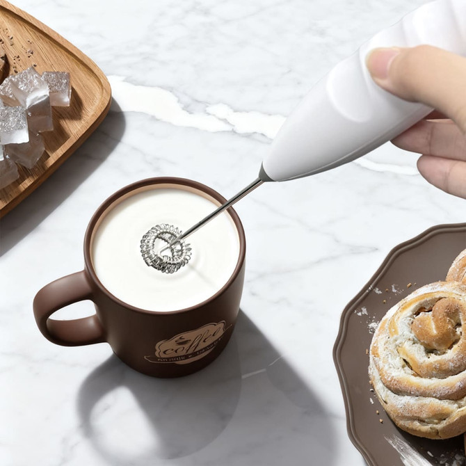 A white milk frother is being used in a brown mug filled with milk, alongside pastries and a wooden tray.