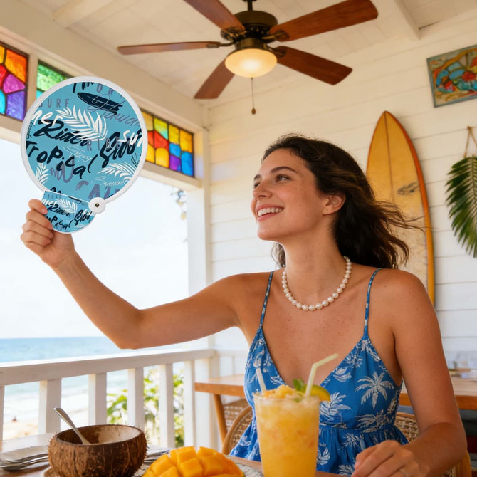 A 20cm foldable reflective fan featuring a tropical design, held by a woman in a blue floral dress, with drinks on the table.