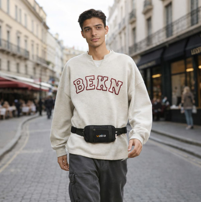A man wearing a light grey sweater walks through a street, sporting a black travel fanny pack around his waist.