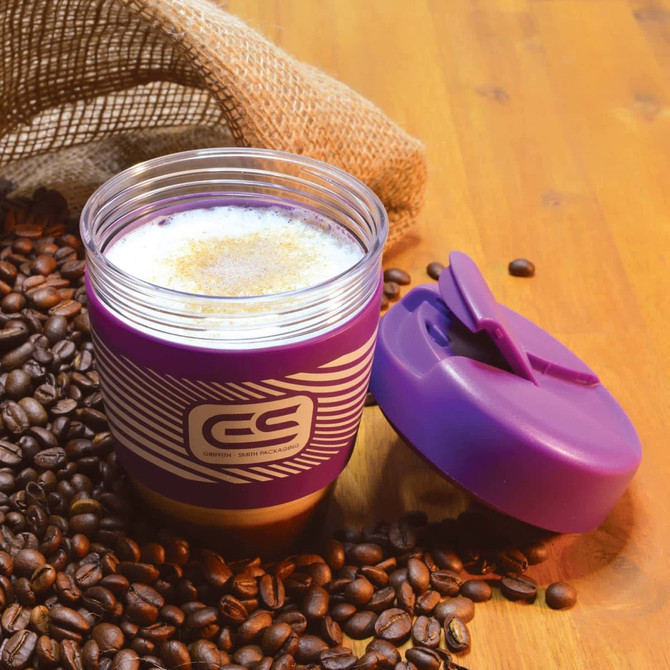 A reusable coffee cup with a purple silicone band and a matching lid, surrounded by coffee beans on a wooden surface.