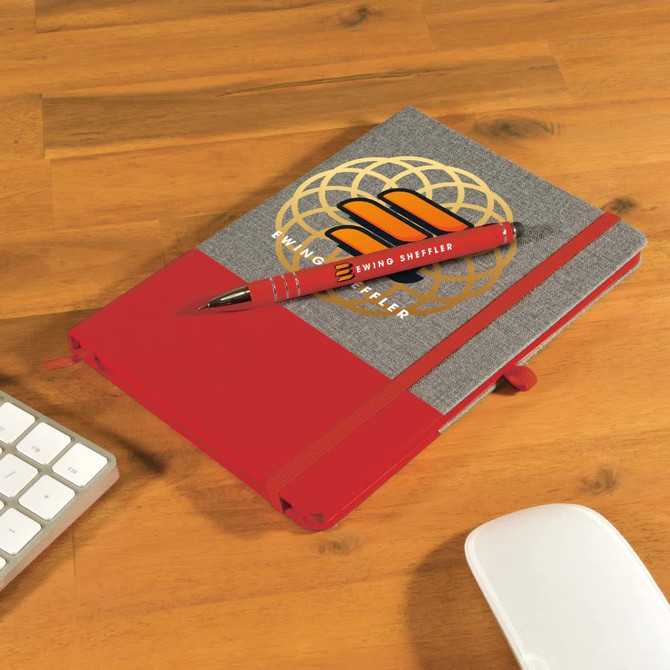 A grey notebook with a red band and a matching red pen placed on a wooden desk alongside a keyboard and mouse.