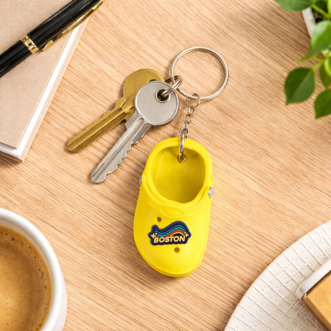 A bright yellow clog shoe keyring is displayed on a wooden surface with keys, a coffee cup, and stationery.
