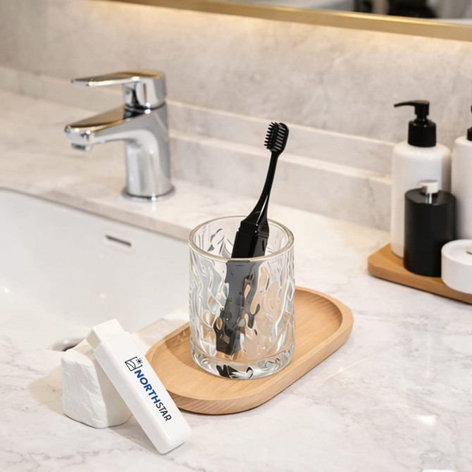 A foldable travel toothbrush in a glass, placed on a wooden tray alongside a bar of soap and bathroom accessories.