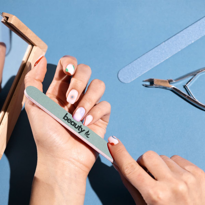 A double-sided polished nail file in green and pink, held in a hand with freshly manicured nails, alongside glasses.