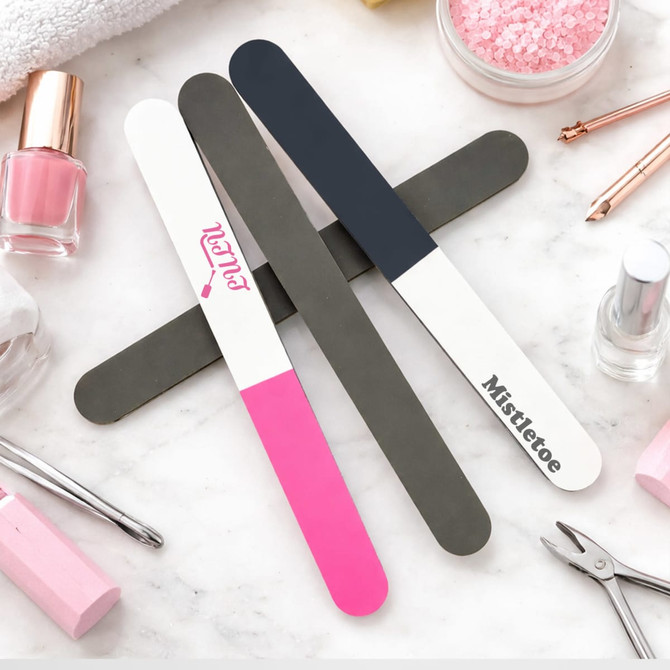 Three triple-sided nail files in pink, grey, and dark blue, arranged on a flat surface with beauty products around.