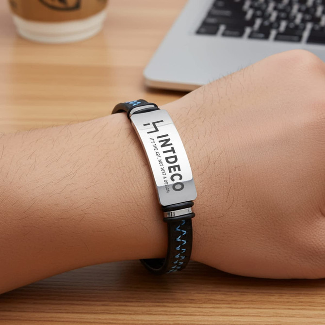 An engraved leather bracelet featuring a silver plate and a black band with blue stitching.
