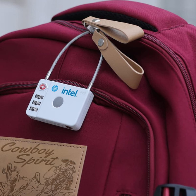 A white luggage lock with a combination dial is attached to a maroon backpack. The lock features a branded logo.