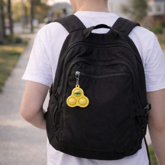 A yellow keychain featuring three bubble buttons hangs from a black backpack. The background includes a path and greenery.
