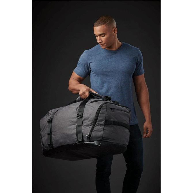 A man holds a large grey duffle bag with straps. The bag features a logo and a sleek design.