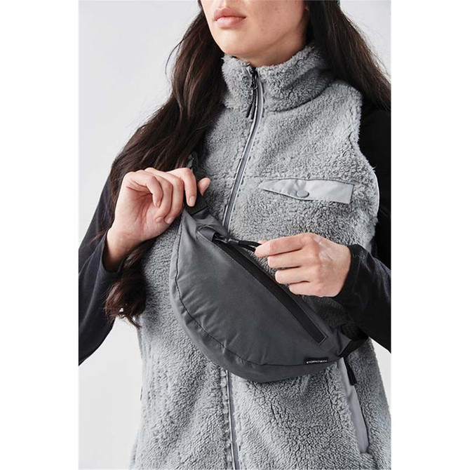 A grey Teton waist pack is being held by a person wearing a textured grey vest. The pack features a zippered compartment and branding.