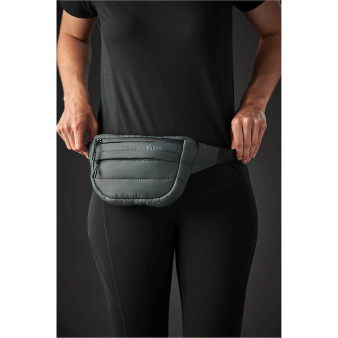 A quilted waist bag in dark grey is being worn around the waist, featuring multiple zippered compartments.