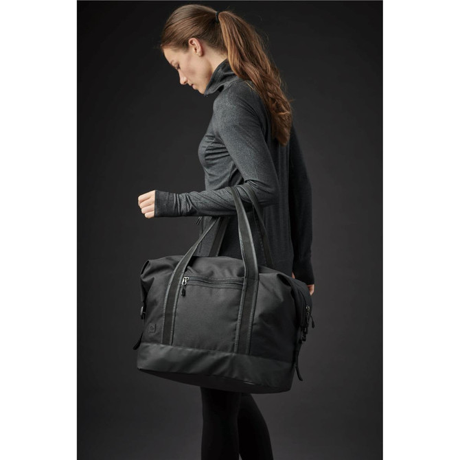 A black duffle bag with dual handles, being held by a woman in athletic attire, against a dark background.