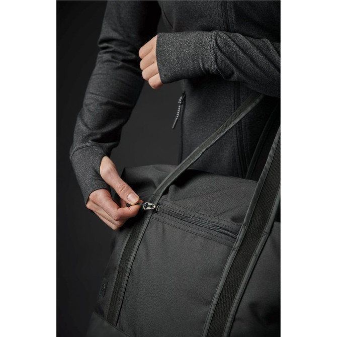 A grey duffle bag being zipped closed, held by a person wearing a dark jacket. The bag features a logo.