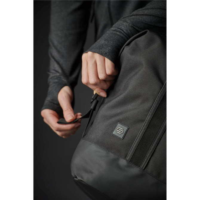 A close-up of a person holding a black duffle bag with a logo, wearing a dark grey long-sleeve top.