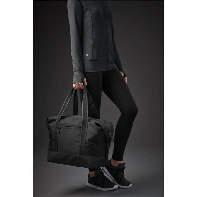 A black duffle bag with sturdy handles, held by a person wearing a dark outfit and sneakers.