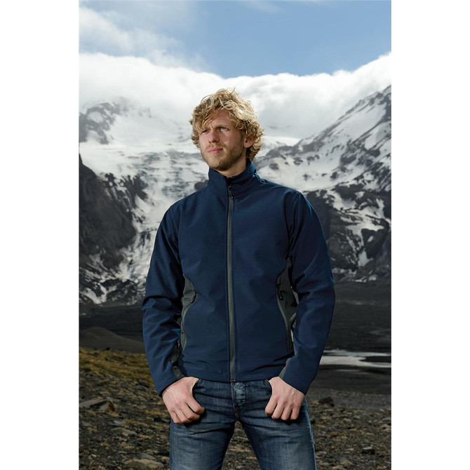 A man wearing a navy softshell jacket stands against a mountainous backdrop, featuring clouds and snow-capped peaks.