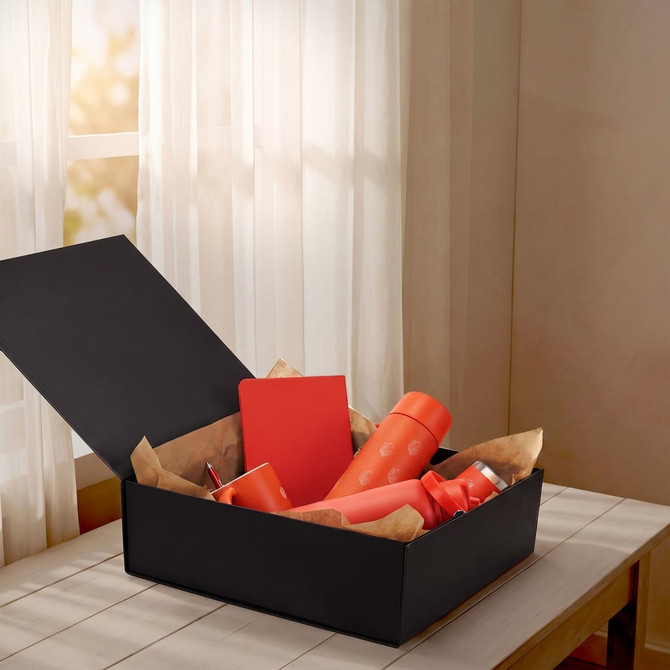 A large black gift box filled with various orange items, including drink bottles and a notebook, resting on a wooden table.