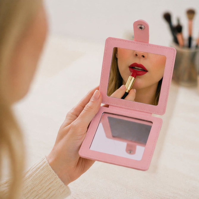 A square folding pocket mirror in pink, reflecting a person applying lipstick. Classic makeup brushes are visible in the background.