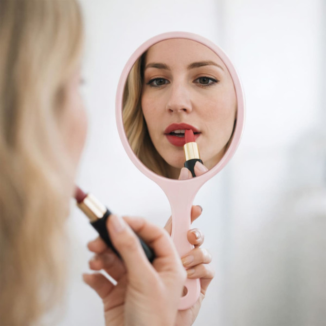 A pink handheld makeup mirror reflects a person applying lipstick, with a soft, neutral background.