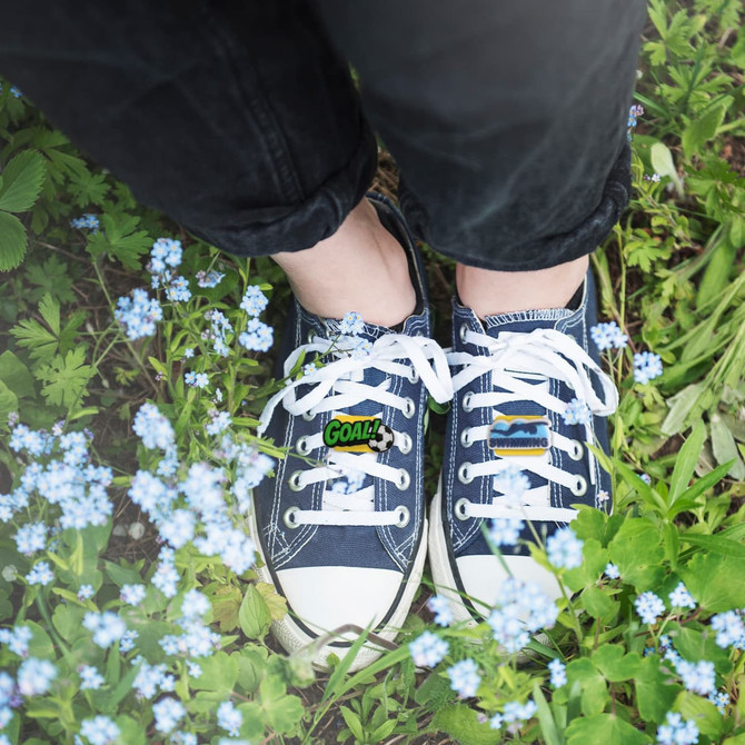 A pair of navy shoes adorned with colourful custom shoe charms, surrounded by small blue flowers and green grass.