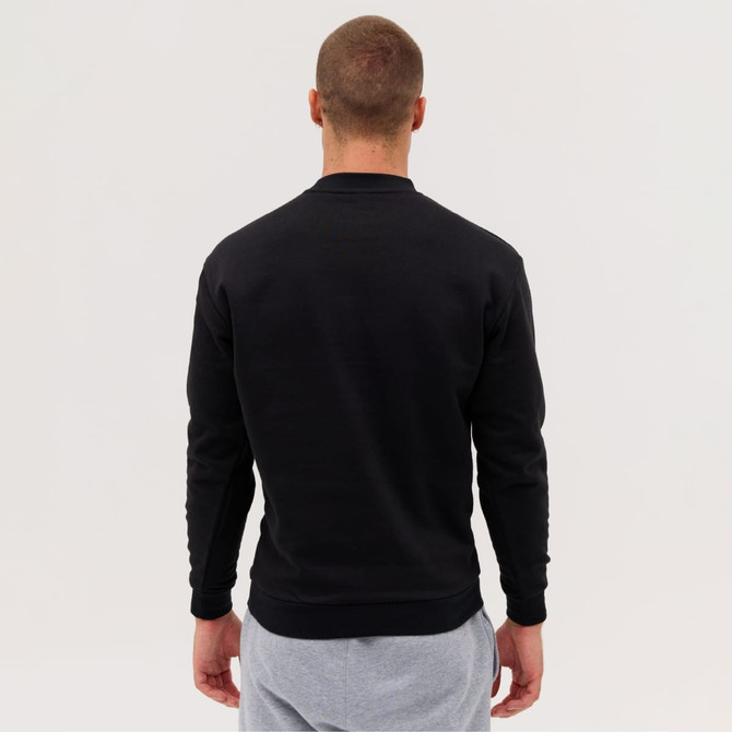 A man is wearing a black sweatshirt, facing away, with light grey trousers. The sweatshirt features a logo.