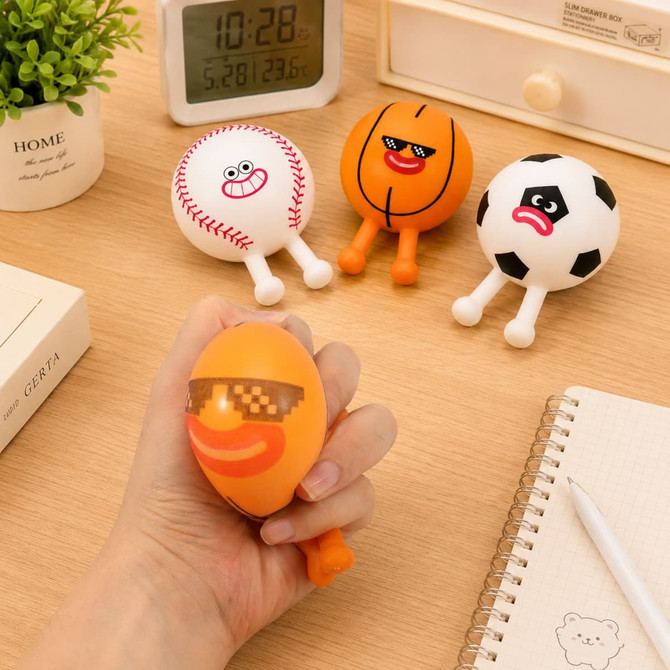 A squishy stress relief ball in orange, resembling a basketball, is held in hand with other sport ball designs nearby.