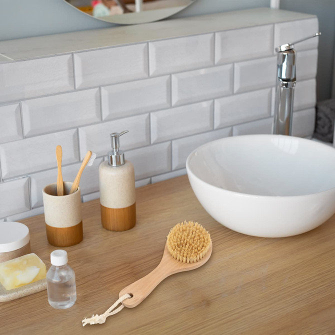 A round body brush with a short wooden handle sits on a wooden countertop near a white bowl and various bathroom items.