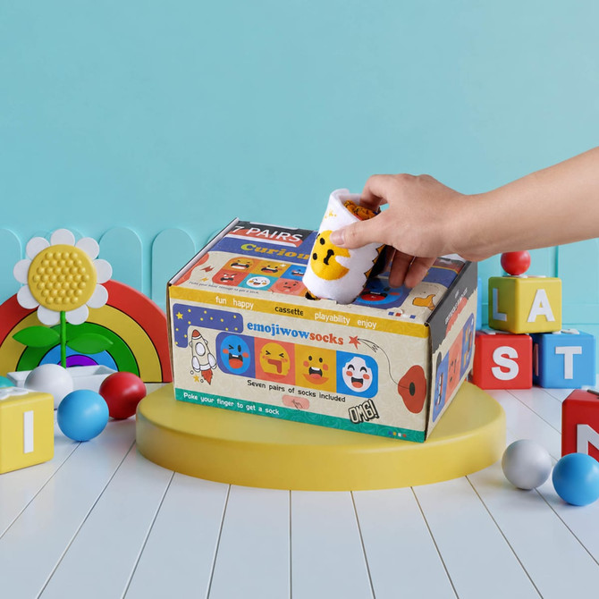 A colourful promotional box featuring various emoji designs, with a hand placing an item inside. The background includes playful toys.