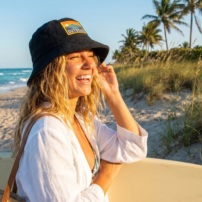 A woman wearing a black terry towelling bucket hat with a full-colour patch, smiling on a beach with palm trees.
