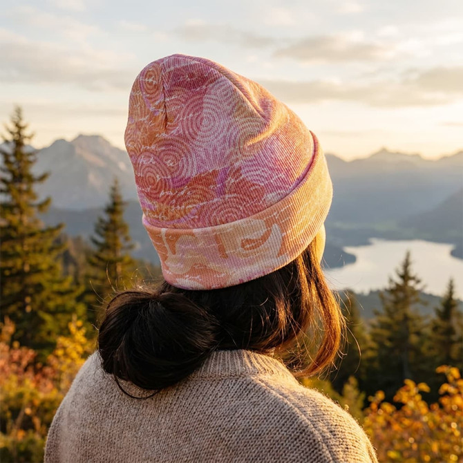 A full-colour beanie in shades of pink and orange, worn by a person against a scenic mountain backdrop.