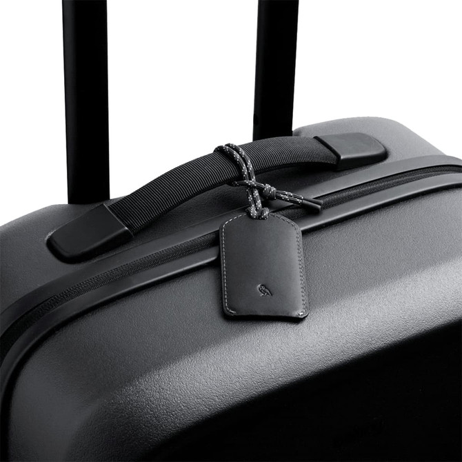 A black suitcase featuring a sleek leather luggage tag with a braided attachment. It has a minimalist design and a logo.