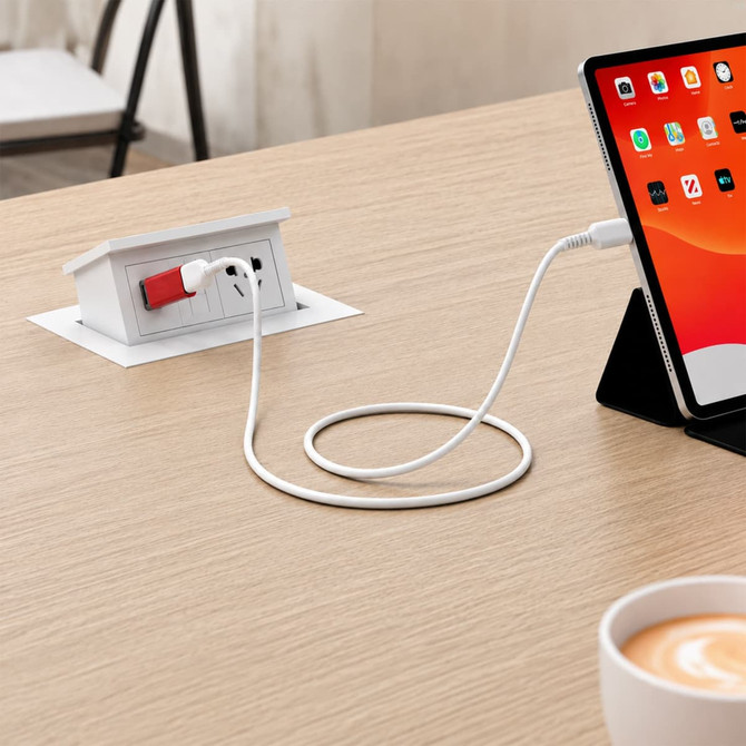 A USB data blocker in white connects to a device beside a tablet on a wooden table with a coffee cup nearby.