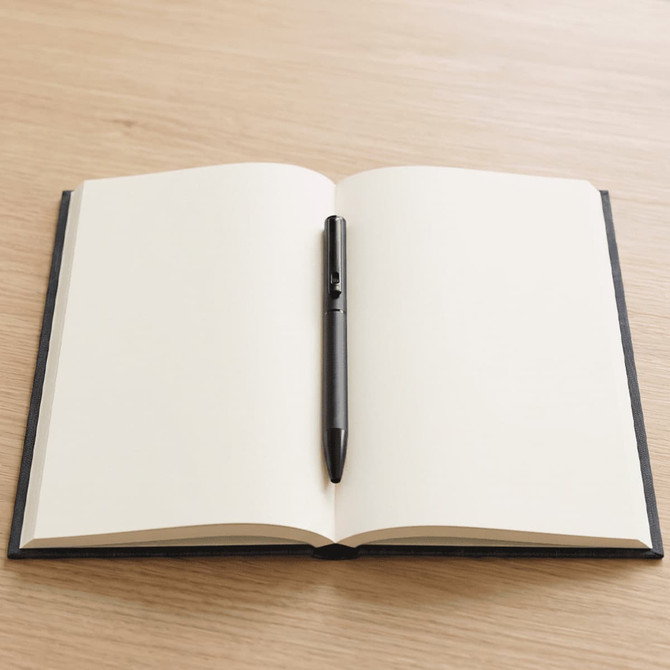 A black ballpoint pen rests on an open, blank, black-covered notebook against a wooden background.