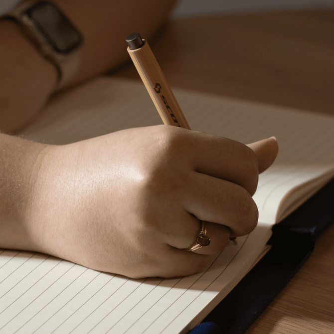 A hand holding a pen writes in a lined, cream-coloured notebook on a wooden surface.