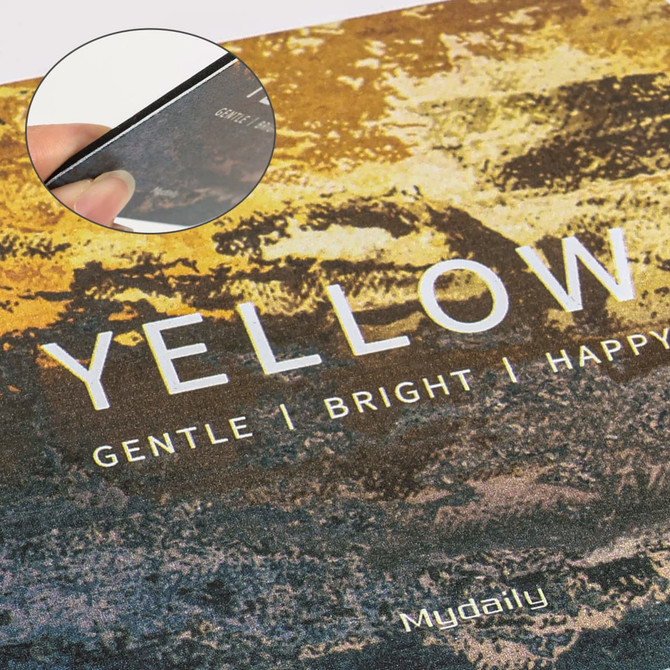 A hand holding the edge of a yellow frosted metal coaster on a textured surface. The coaster has a logo.