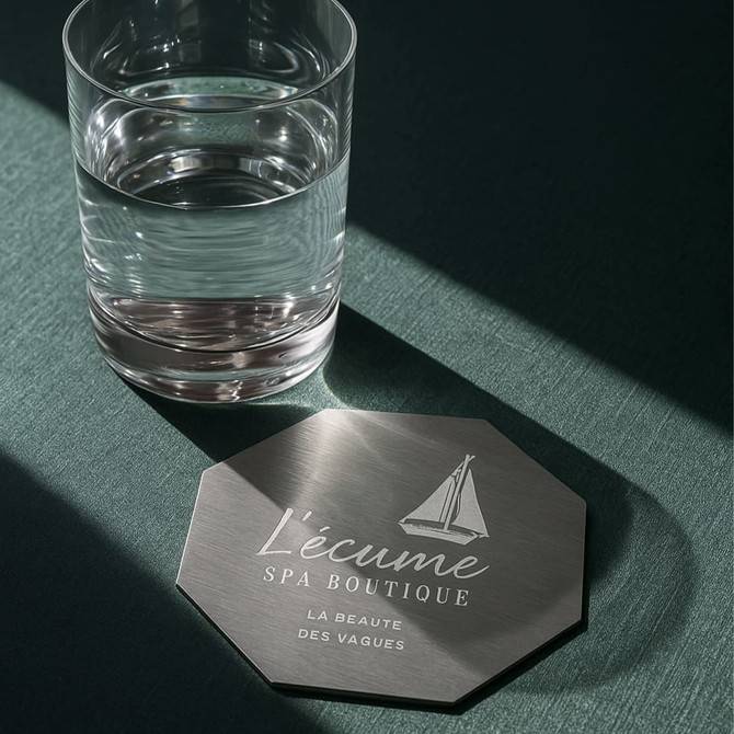 An octagonal metal coaster with a silver finish rests on a green surface beside a glass of water.