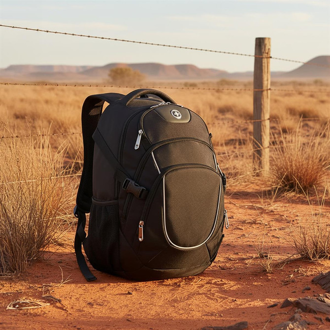 A black backpack with zippers and a logo rests on sandy terrain, surrounded by dry grass and a fence.