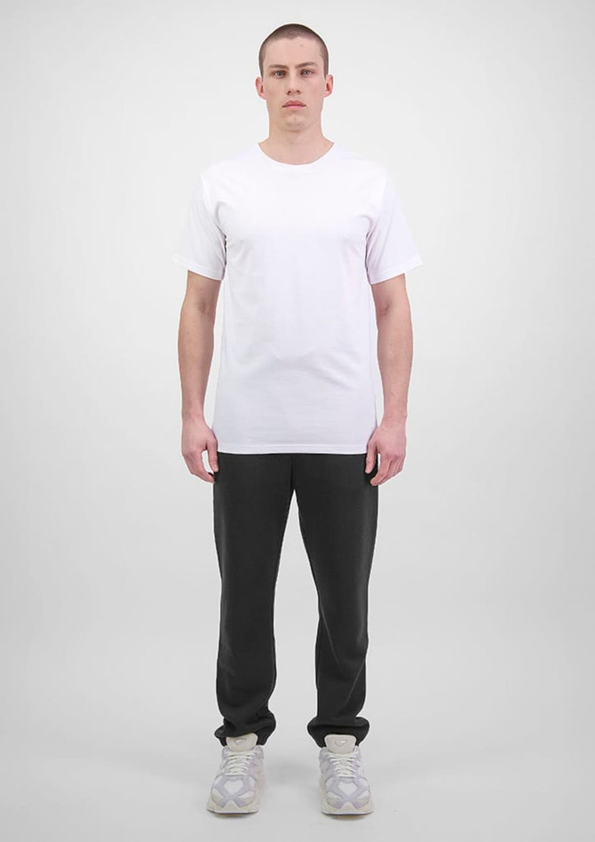 A man stands wearing a plain white t-shirt and black track pants. The track pants feature a logo.